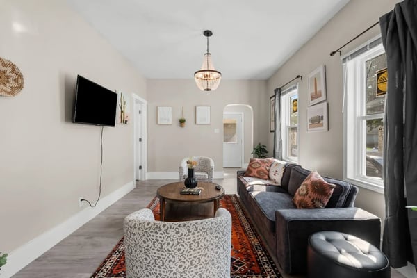 Modern living room with gray sofa, patterned armchairs, round wood table, wall-mounted TV, pendant light, and large windows with gray curtains