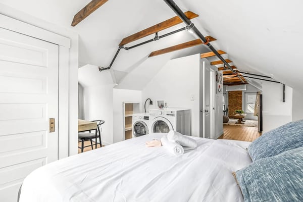 Attic bedroom with exposed wooden beams, white walls, bed in foreground, and washer-dryer visible in background