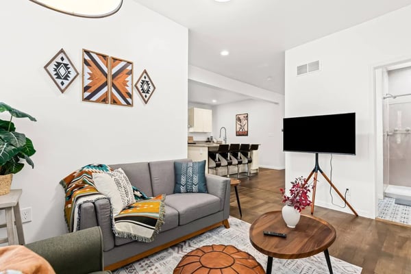 Modern living room with gray sofa, geometric wall art, wooden coffee table, TV on stand, and open kitchen in background