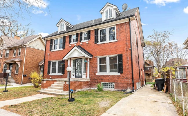 Two-story red brick house with two dormers, white trim, black shutters, and front porch in residential neighborhood.
